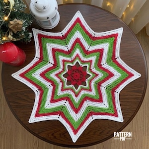 Op de afbeelding: Een stervormig gehaakt kleedje met concentrische ringen van wit, groen en rood garen. Het midden bestaat uit een klein, ingewikkeld rood bloemmotief, omringd door afwisselende puntige lagen van witte, groene en rode steken.