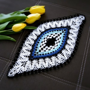Op de afbeelding: Een handgemaakte, oogvormige doily in wit, blauw en zwart. Het haakontwerp heeft een centrale zwarte cirkel, omgeven door blauwe en witte ringen. De doily is afgezet met een zwarte rand. Gele tulpen staan op de achtergrond.