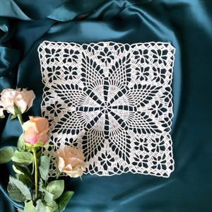 May include: A white crocheted square doily with a delicate floral pattern. The doily is laid on a teal fabric background.