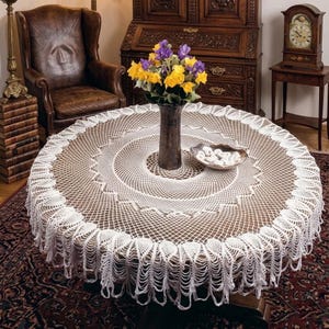 May include: A round, white crocheted tablecloth with a detailed, openwork design. The tablecloth is draped over a wooden table, and the edge features a scalloped fringe. A vase of yellow and purple flowers sits in the center.