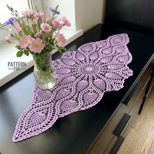May include: A light purple crochet doily with an intricate, elongated floral-inspired pattern featuring a central circular motif and radiating leaf-like shapes. The doily is displayed alongside a clear glass vase filled with pink and blue flowers.