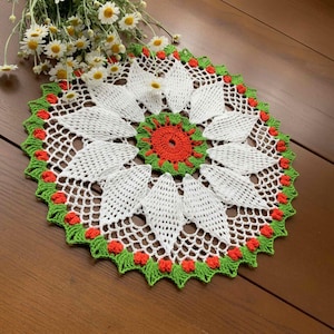 May include: A handmade, white crochet doily with a floral design. The center is orange and green, surrounded by white petals and a green and red scalloped edge. Daisies are placed on the top left of the doily.