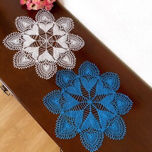 May include: Two crocheted doilies, one white and one blue, with a delicate, intricate design. The doilies are laid out on a wooden surface.