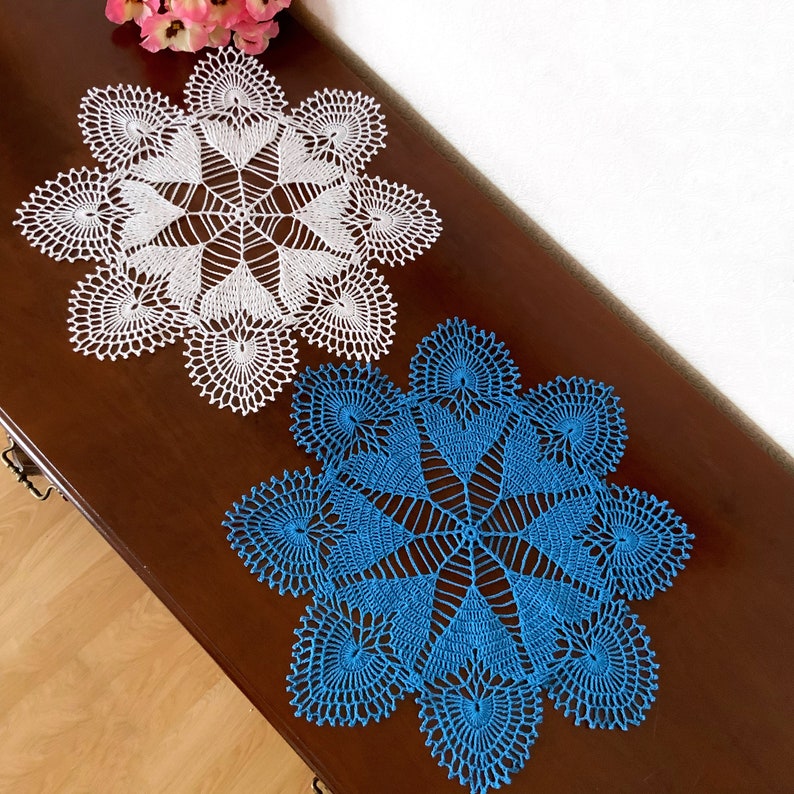 May include: Two crocheted doilies, one white and one blue, with a delicate, intricate design. The doilies are laid out on a wooden surface.