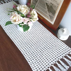 May include: A white, open-weave crochet table runner with a fringed edge. The runner is displayed on a dark wood surface, with a vase of cream-colored roses and a framed picture in the background.