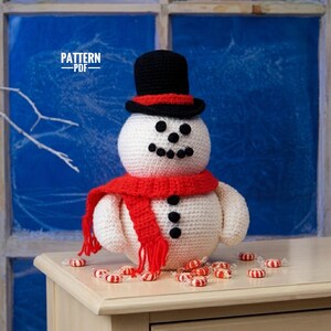May include: A white crocheted snowman featuring a black top hat with a red band, a red scarf with fringe, and black buttons for eyes, mouth, and torso. The text PATTERN PDF is visible in the upper left corner of the image.