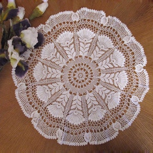 May include: A white, circular, crocheted doily with a detailed floral pattern. The doily is placed on a brown wooden surface, with a bouquet of purple and white irises to the left. The doily's intricate design is visible.