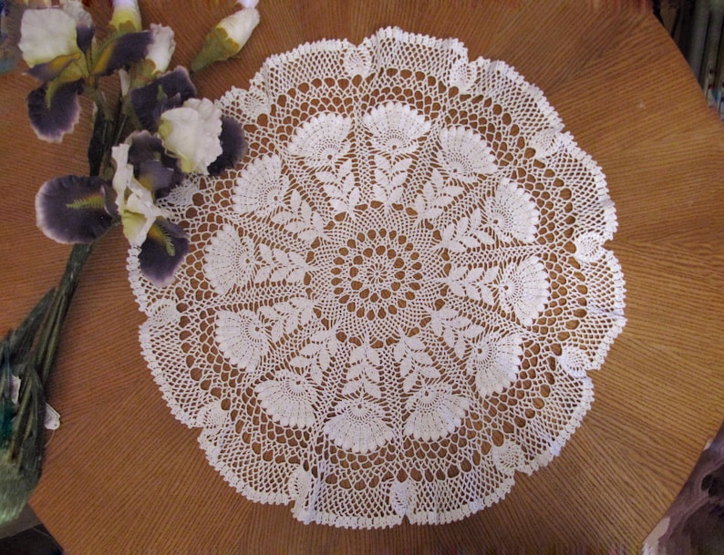 May include: A white, circular, crocheted doily with a detailed floral pattern. The doily is placed on a brown wooden surface, with a bouquet of purple and white irises to the left. The doily's intricate design is visible.