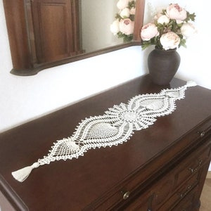 Może przedstawiać: Biały, szydełkowy bieżnik stołowy o delikatnym, skomplikowanym wzorze. Bieżnik ma po jednej frędzli na każdym końcu i leży na ciemnym drewnianym blacie.