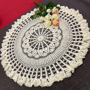 May include: Cream-colored, crocheted doily with a detailed floral pattern. The round doily features an intricate openwork design and a ruffled edge. A bouquet of roses in shades of white and peach rests on the doily.