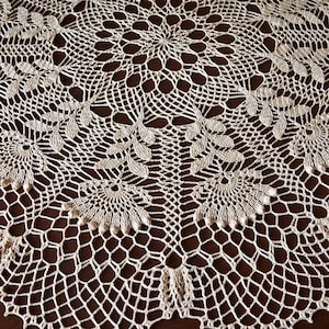 May include: A cream-coloured, handmade crochet doily with an intricate floral pattern. The design features circular and leaf-like motifs, creating a delicate, textured surface. The doily is set against a dark brown background, highlighting the detailed craftsmanship.