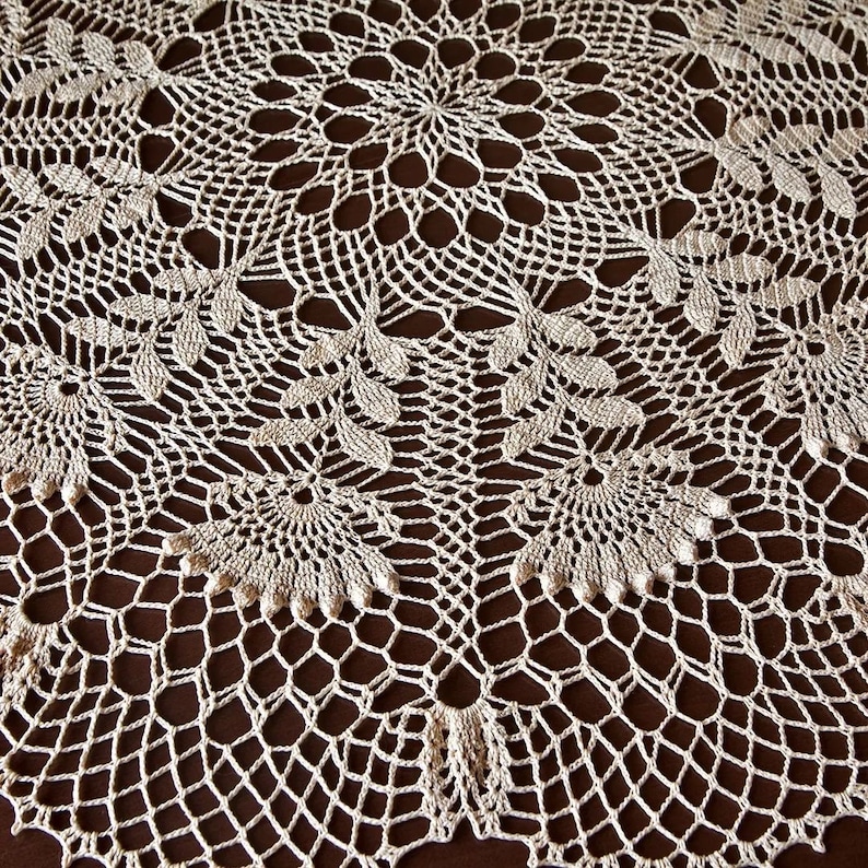 May include: A cream-coloured, handmade crochet doily with an intricate floral pattern. The design features circular and leaf-like motifs, creating a delicate, textured surface. The doily is set against a dark brown background, highlighting the detailed craftsmanship.
