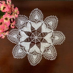 May include: White crocheted doily with a delicate, intricate design. The doily has a circular shape with a starburst pattern in the center and a scalloped edge.