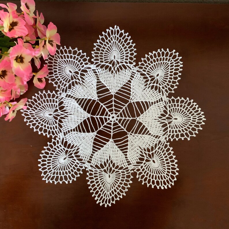 May include: White crocheted doily with a delicate, intricate design. The doily has a circular shape with a starburst pattern in the center and a scalloped edge.