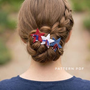 May include: Three crocheted star hair accessories in red, white, and blue, each embellished with small beads. The red star features blue beads, the white star has red and blue beads, and the blue star has red beads. The text PATTERN PDF is visible.