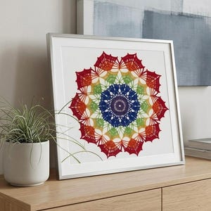 Może przedstawiać: Oprawiony wydruk artystyczny mandali z kolorowym wzorem. Grafika przedstawia symetryczny wzór z koncentrycznymi okręgami w kolorach czerwonym, pomarańczowym, żółtym, zielonym, niebieskim i fioletowym. Wydruk jest w srebrnej ramie, na drewnianej powierzchni.