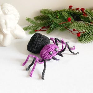 May include: A handmade crochet spider with a purple body, black legs, and a black egg-shaped object. The spider has silver-colored eyes. The background includes a white statue and a garland of evergreen branches with red berries.