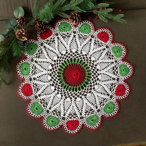 May include: A handmade, white crochet doily with a festive Christmas theme. The doily features a central red and green circle design, surrounded by alternating red and green accents. The intricate lace pattern adds a delicate touch to this holiday decor.