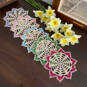 May include: A set of six crocheted doilies in various colours, including pink, blue, green, and white. The doilies are arranged in a fan shape on a wooden surface.