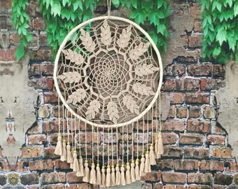 Peut inclure: Un attrape-rêves en crochet beige avec un motif circulaire et des franges. L'attrape-rêves est suspendu à un mur de briques avec des vignes vertes.