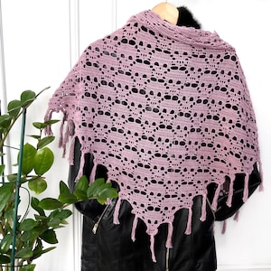 May include: A mauve-colored, open-weave crochet shawl with a skull pattern. The shawl has a fringed edge and is draped over a black jacket. The shawl is made of a soft yarn and is a unique accessory.