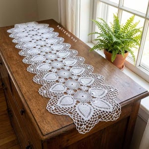 May include: A white rectangular crochet table runner featuring a repeating intricate floral and leaf pattern with a scalloped edge. The textile is displayed flat, and the text PATTERN PDF is visible on the surface of the fabric.