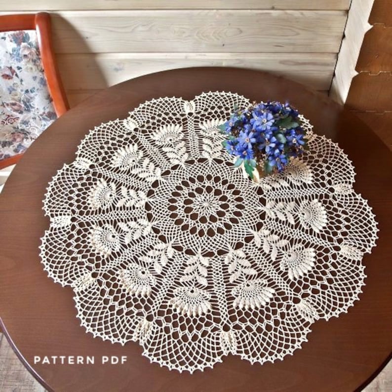 May include: Cream-coloured, crocheted doily with a floral design, resting on a dark brown wooden table. The doily features intricate patterns and a scalloped edge. A small bouquet of blue flowers sits on top. The text "PATTERN PDF" is visible.