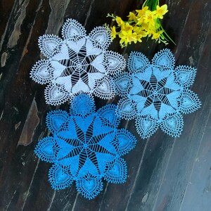 May include: Three crocheted doilies, one white and two blue, with intricate patterns. The doilies are arranged on a dark wooden surface.