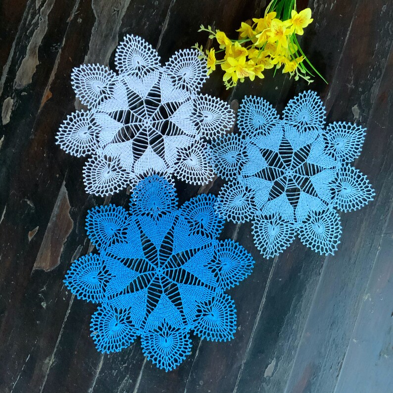 May include: Three crocheted doilies, one white and two blue, with intricate patterns. The doilies are arranged on a dark wooden surface.