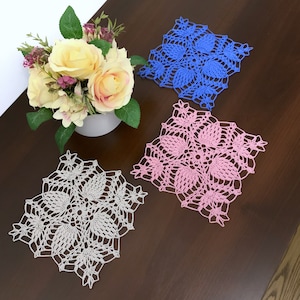 May include: Three crocheted doilies in different colors: white, pink, and blue. The doilies are square shaped and have a delicate floral design.