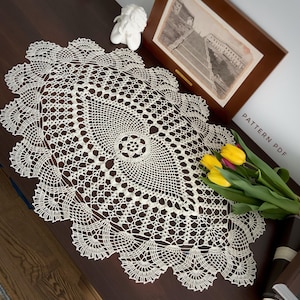 May include: A white crocheted doily with a delicate floral pattern. The doily is laid out on a wooden surface with a bouquet of yellow flowers and a book.