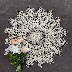 May include: A delicate, off-white crocheted doily with a swirling pattern in the center. The doily is laid on a dark gray surface with a bouquet of white and pink flowers.