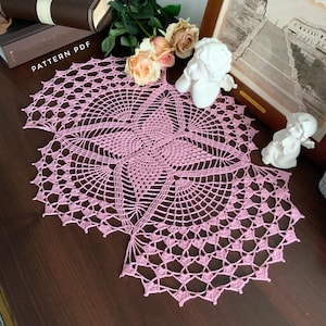 Peut inclure: Un napperon en crochet rose avec un motif de papillon. Le napperon est posé sur une surface en bois foncé.