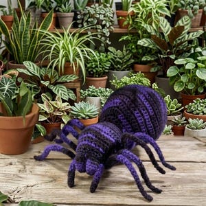 May include: A handmade crochet spider in shades of purple and black, with eight long legs and a round body. The spider is placed on a wooden surface, with a variety of potted plants in the background.