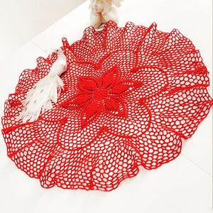 Op de afbeelding: Rood gehaakt tafellaken met een bloemmotief in het midden. Het tafellaken is rond en heeft een gegolfde rand.