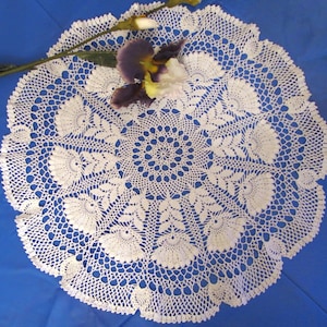 May include: A white, circular, crocheted doily with a floral design. The doily features a central circular pattern surrounded by detailed floral and leaf motifs. A purple and yellow flower rests on the doily.