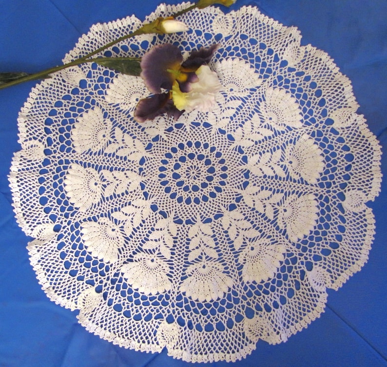 May include: A white, circular, crocheted doily with a floral design. The doily features a central circular pattern surrounded by detailed floral and leaf motifs. A purple and yellow flower rests on the doily.