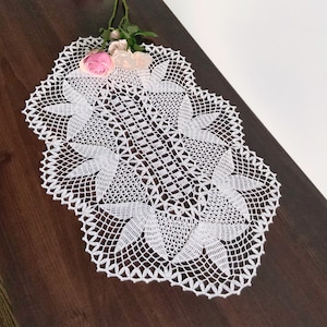 May include: White crocheted doily with a delicate floral pattern. The doily is oval shaped and has a scalloped edge.