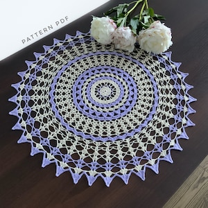Peut inclure: Un napperon crocheté avec un motif violet et blanc. Le napperon est rond et présente un motif de dentelle délicat. Il est posé sur une table en bois.
