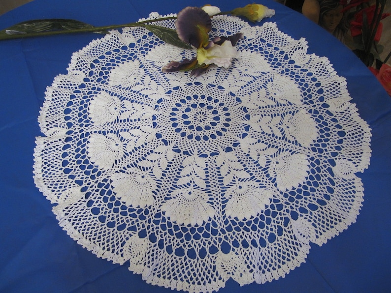 May include: A white, circular, crocheted doily with a floral pattern. The doily is on a blue surface and is decorated with a purple and yellow flower.