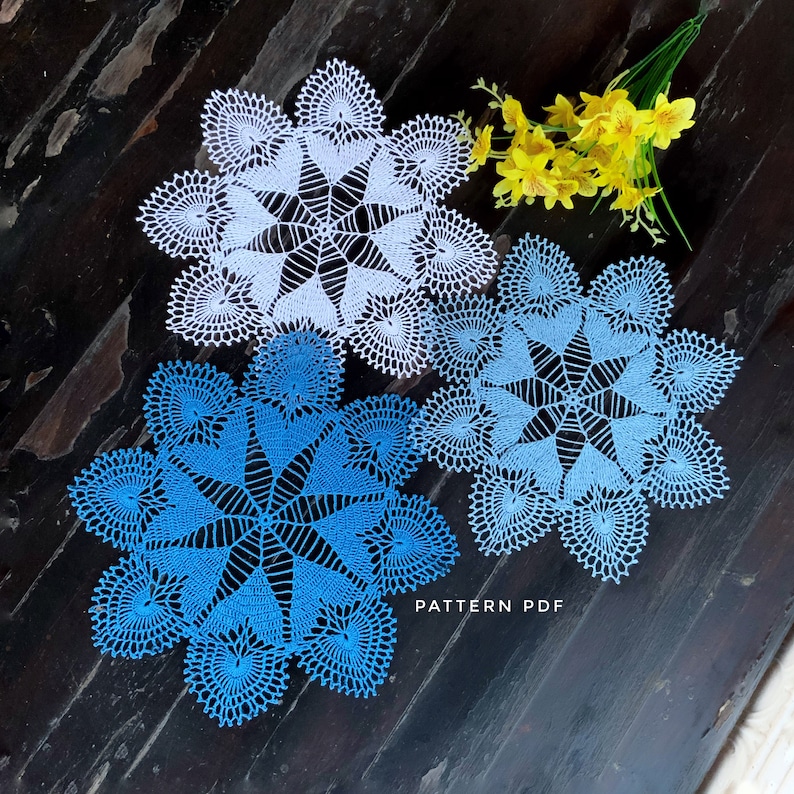 May include: Two crocheted doilies, one white and one blue, with a delicate, intricate design. The doilies are laid out on a dark wooden surface with a bouquet of yellow flowers.