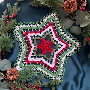 May include: A white, green, and red crocheted star with a detailed design. The star is on a dark blue fabric background.