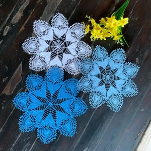 May include: Two crocheted doilies, one blue and one white, with a delicate floral pattern. The doilies are laid out on a dark wooden surface.