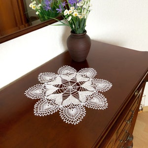 May include: White crocheted doily with a delicate floral design, placed on a dark wood surface with a vase of purple and white flowers.