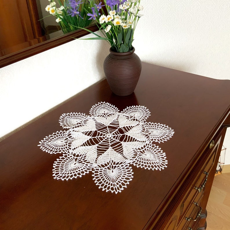 May include: White crocheted doily with a delicate floral design, placed on a dark wood surface with a vase of purple and white flowers.