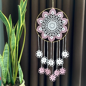 May include: A handmade dreamcatcher with a circular design. The dreamcatcher features a white and pink floral crochet pattern within a gold-colored hoop. White strings hang down, adorned with white and pink crochet flowers. The background is a brown curtain.
