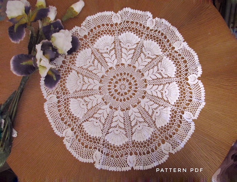 May include: A white, circular, crocheted doily with a detailed floral pattern. The doily is placed on a brown wooden surface. The text "PATTERN PDF" is visible in the lower right corner. The doily is a handmade craft item.