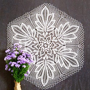 Könnte beinhalten: Weißes, sechseckiges Deckchen mit aufwendigem Lochmuster. Das Deckchen liegt auf einer dunklen Oberfläche, mit einem Strauß lila Blumen links. Die Blumen sind in braunes Papier gewickelt.