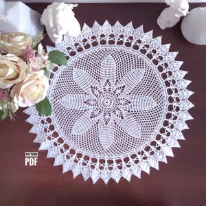 以下が含まれることがあります： 中央に花の模様、縁にスカラップ模様が施された白いかぎ針編みのドイリー。 ドイリーは円形で、濃い茶色の表面に置かれ、背景には花や装飾的な要素があります。 テキスト「PATTERN PDF」が表示されています。