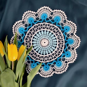 Op de afbeelding: Een handgemaakte, wit en blauw gehaakte doily met een cirkelvormig ontwerp. De doily heeft een centraal patroon van witte en lichtblauwe concentrische cirkels, omgeven door een gedetailleerde, opengewerkte rand. Gele tulpen staan in de linker benedenhoek.